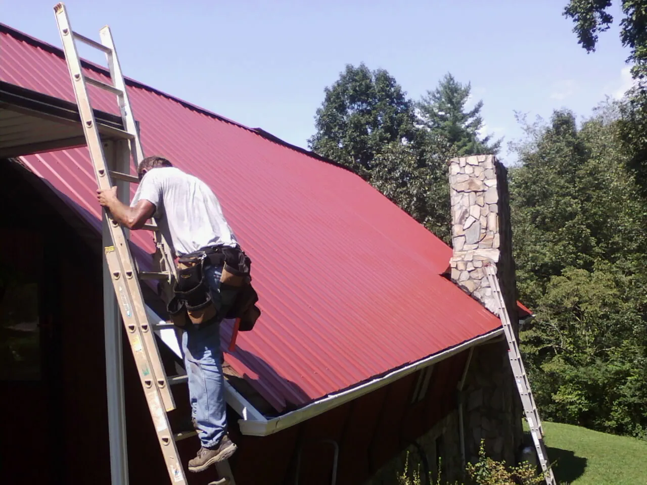 Roofer on a ladder beside a red metal roof and stone chimney — Soffit & Fascia Repair by Roofing Brothers in Piney Woods